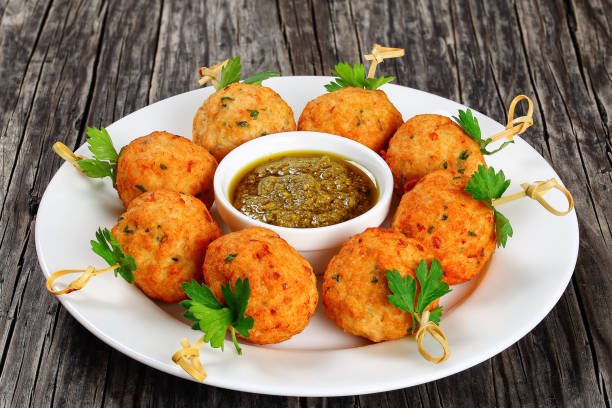 delicious roasted meatballs on wooden skewers with parsley on white plate with homemade basil, parmesan olive oil sauce in gravy boat on old dark wooden table, horizontal view from above, close-up